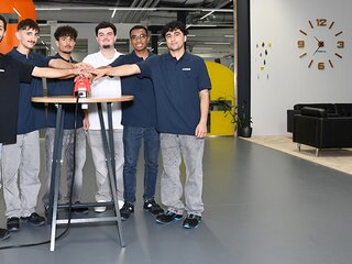 Ausbildungsstart 2025 Sechs junge Menschen stehen in Arbeitskleidung um einen Tisch und drücken gemeinsam einen roten Knopf, Symbol für den Start einer Ausbildung.